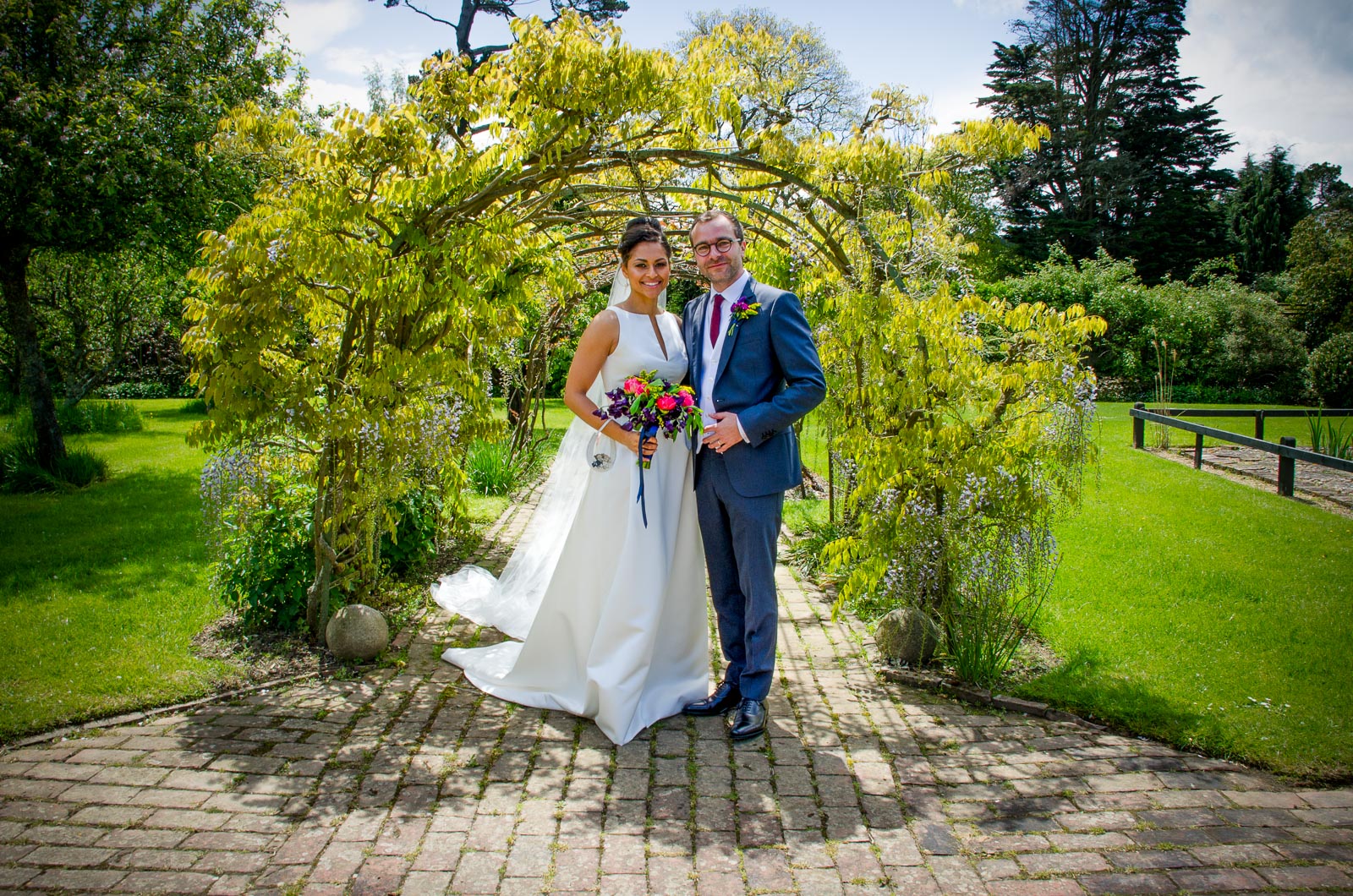 Tash and Raf pose in the gardens in Borde Hill, Haywards Heath under a leafy arch after their wedding.