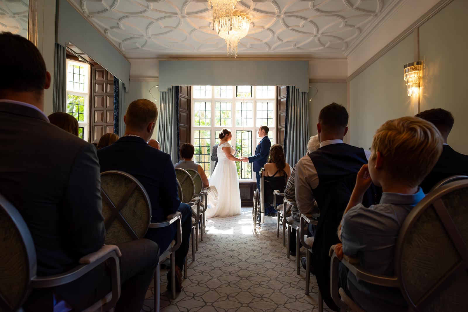 Amy and James hold hands surrounded by wedding guests in the Ainsworth Room in Lewes Register Office during their wedding.