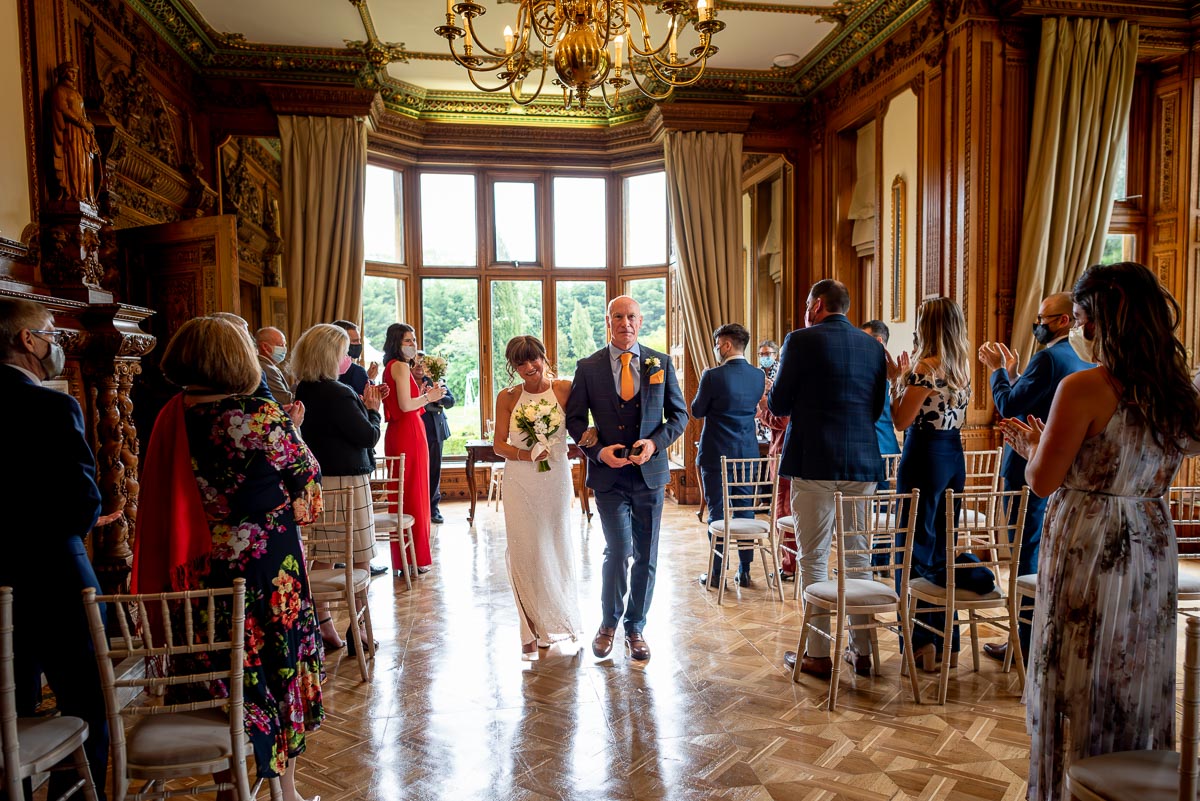 Carmen and Jeff walk towards the camera amongst seated guests in Maximilian, a room in Manor by the Lake, Cheltenham.