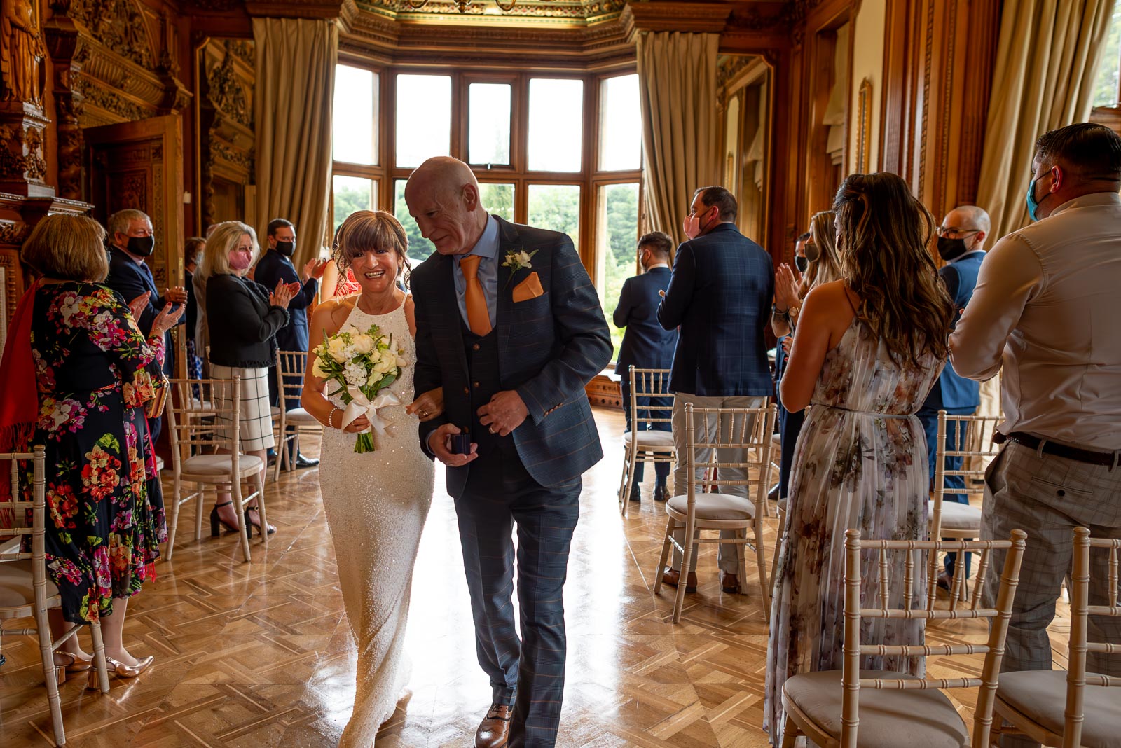 Carmen and Jeff walk towards the camera amongst seated guests in Maximilian, a room in Manor by the Lake, Cheltenham.