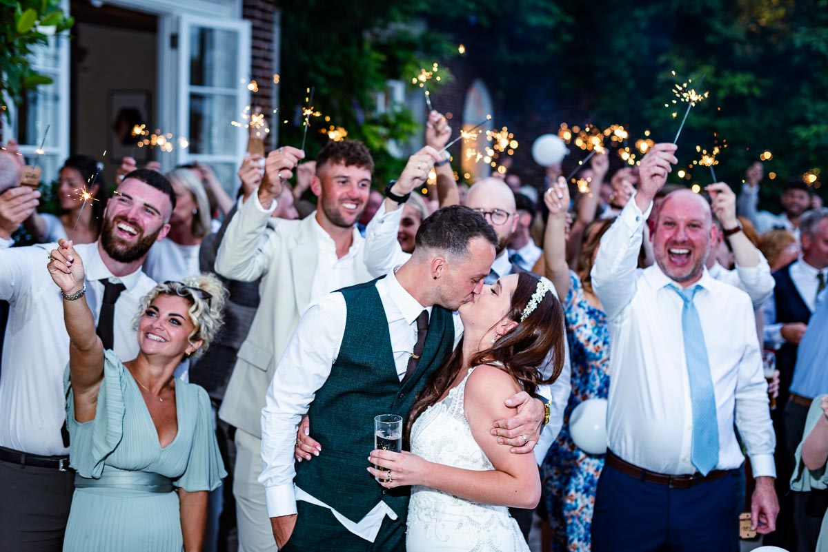 Natalie and Dean embrace at the rear of Pelham House Hotel, Lewes after their wedding with friends and family holding sparklers and cheering in the background.