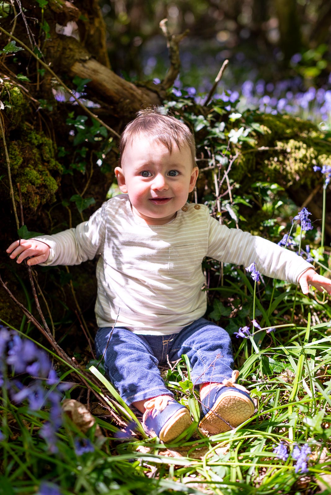 Eight month old baby Alasdair sits down and smiles at the camera among the bluebells at Battle Great Woods.