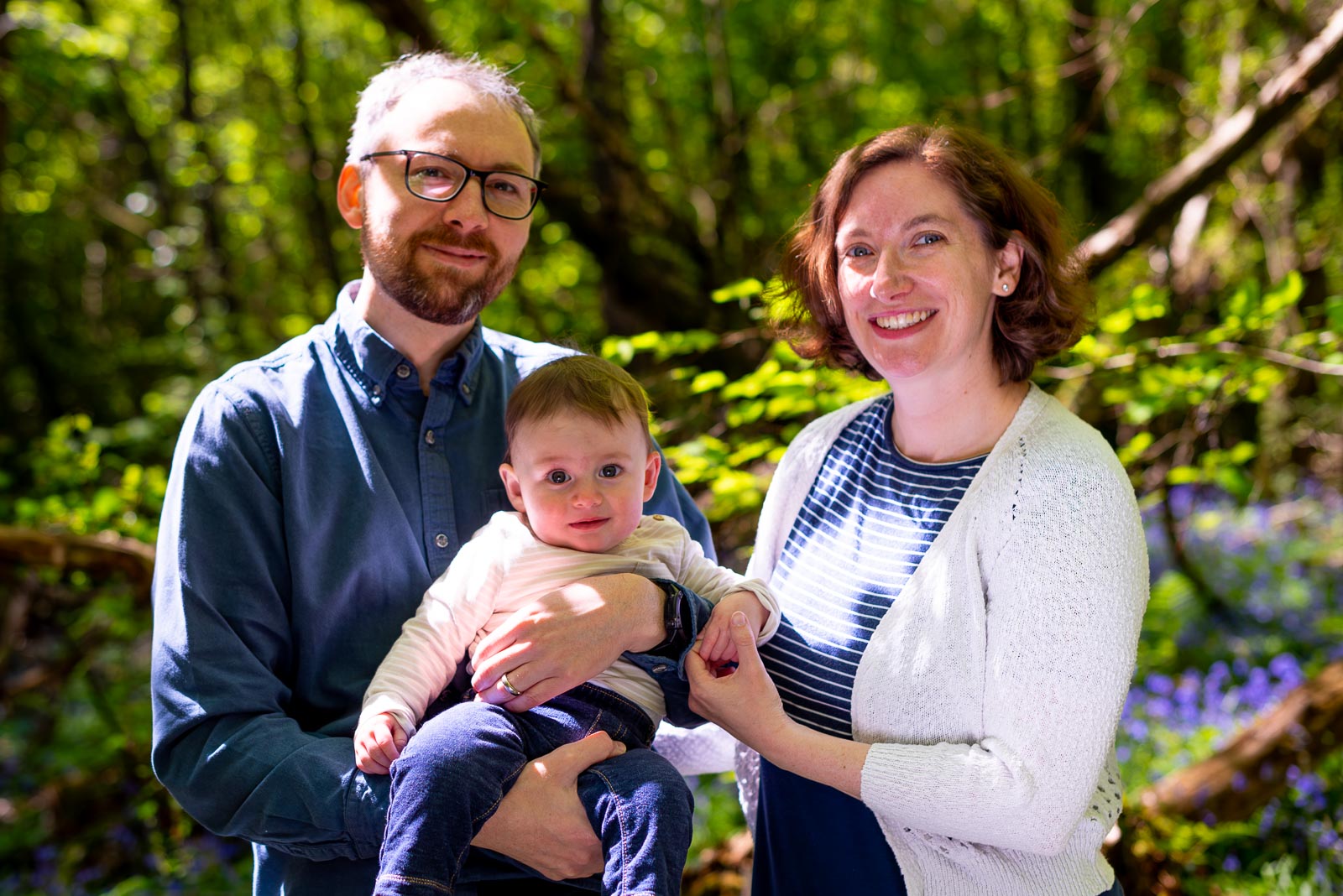 Alexis, Niell and their eight month old baby Alasdair smile at the camera among the bluebells at Battle Great Woods.