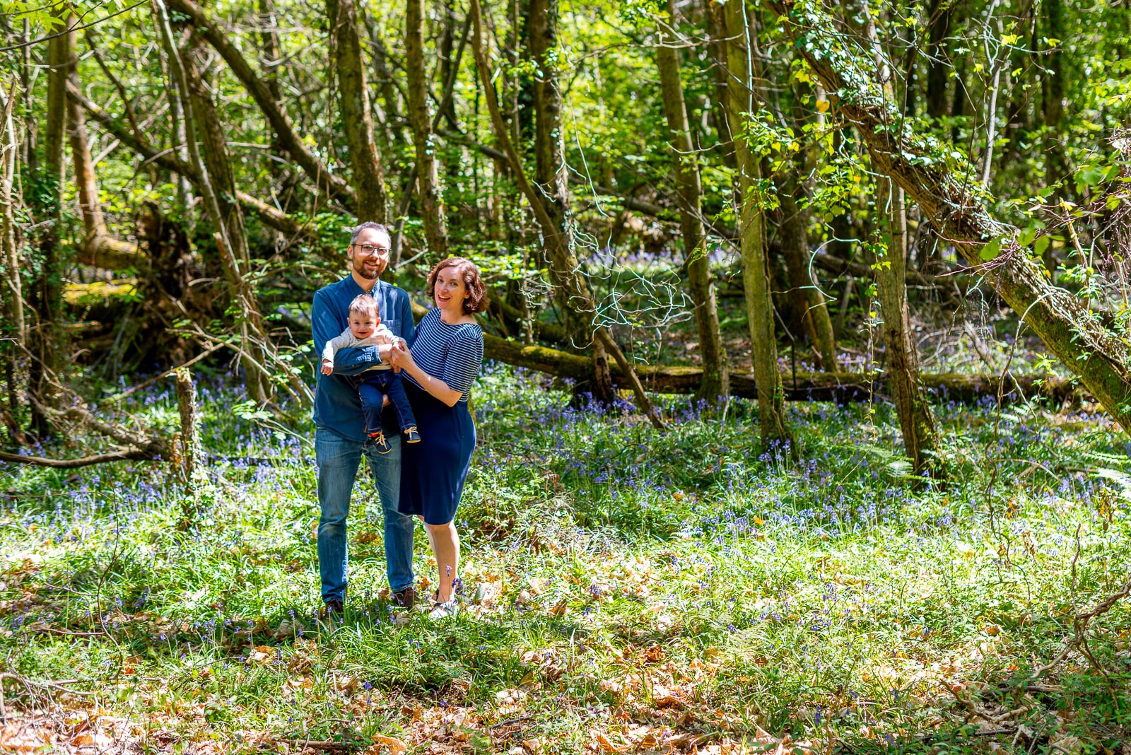 Neill, Alexis and their eight month old baby Alasdair smile at the camera in a wide angle photograph among the bluebells at Battle Great Woods.