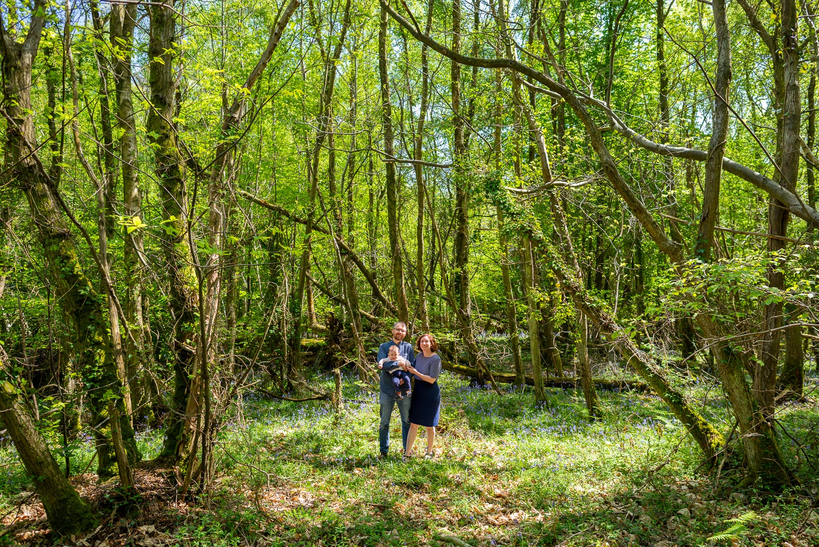 Neill, Alexis and their eight month old baby Alasdair smile at the camera in a super wide angle photograph among the bluebells at Battle Great Woods.