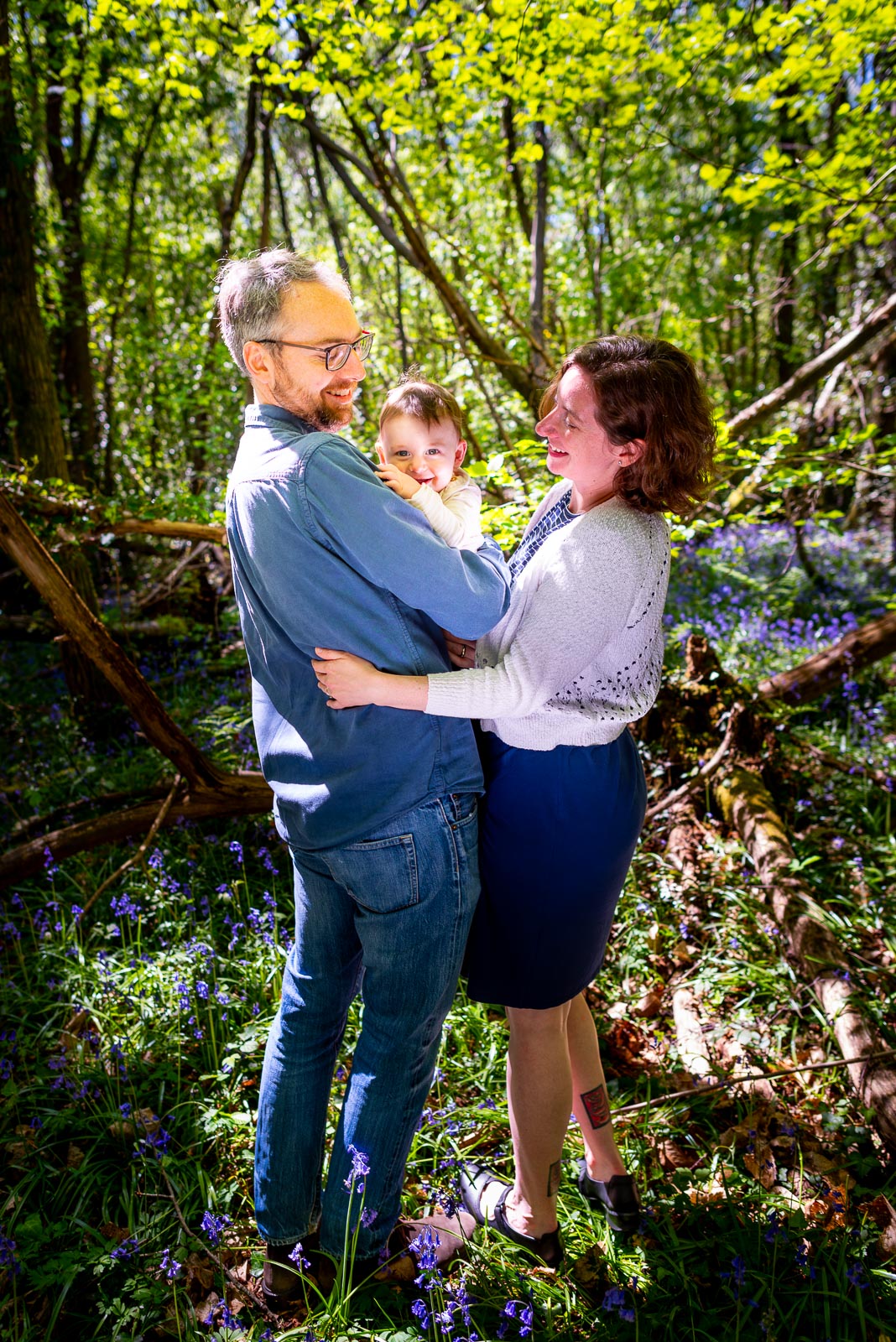 Alexis and Niell smile at their eight month old baby Alasdair who smiles over Niell's shoulder among the bluebells at Battle Great Woods.