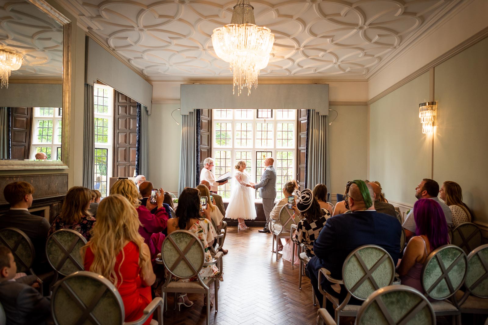 Lou and Matt are surrounded by their seated friends and family whilst standing at the top of the aisle in the Ainsworth Room at Lewes Register Office.