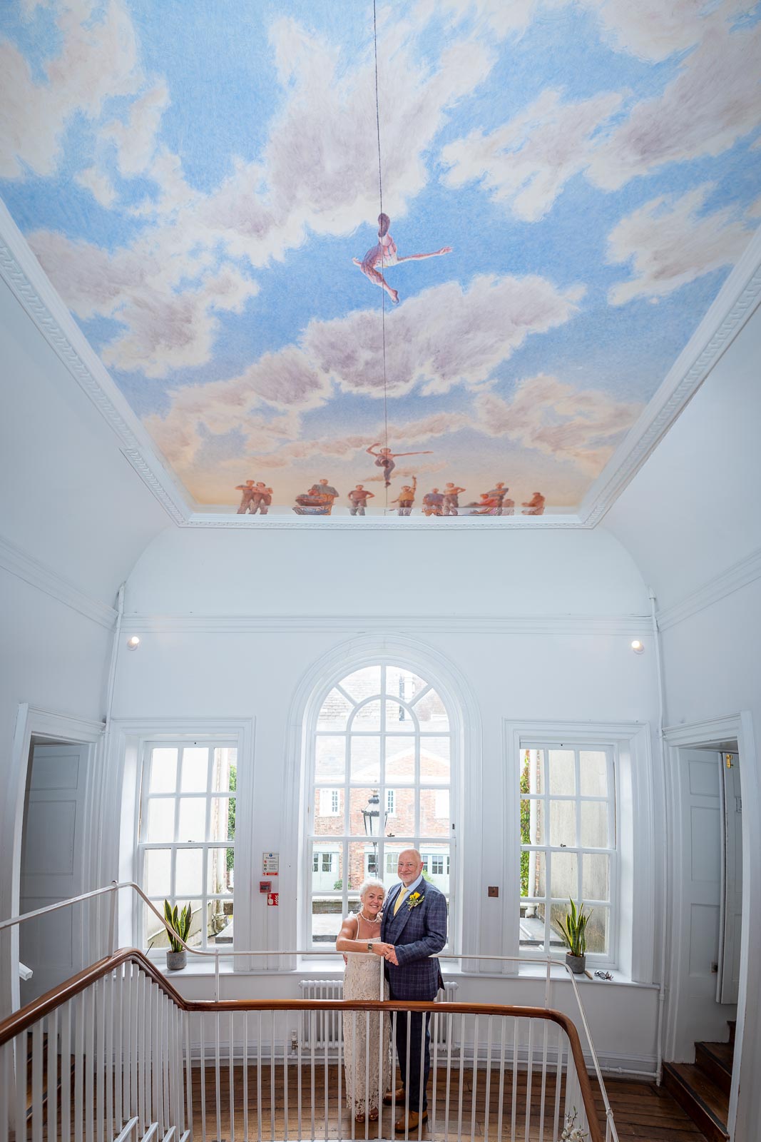 Karen and Hugh smile on the stairs of Pelham House in Lewes underneath the tight rope roof painting following their wedding. 