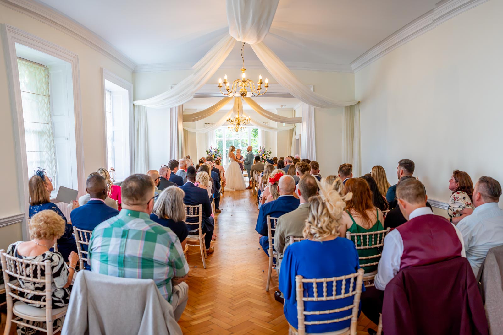 Sarah and Lewis surrounded by guests during their wedding at the bottom of the aisle in the Goring Room at Pelham House Hotel, Lewes. 