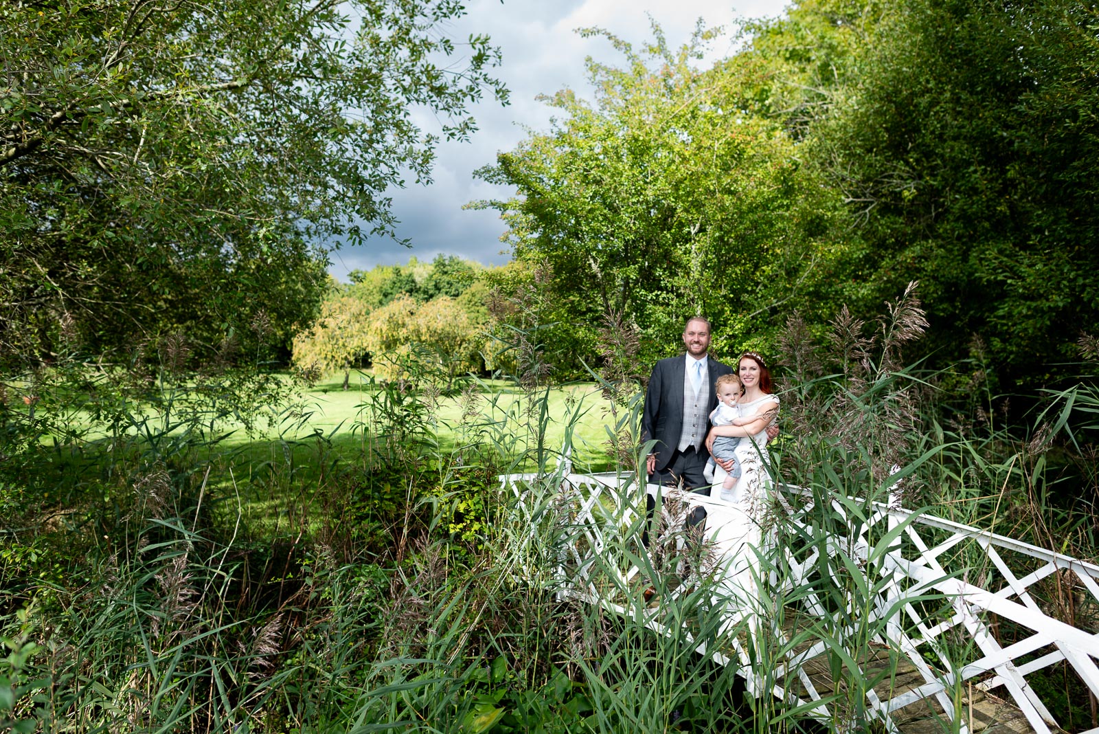 Adrienn, Lee and their youngest son pose on the white lattace bridge at Milwards Farm Estate near Lewes after their wedding